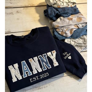 May include: A navy blue sweatshirt embroidered with the word "NANNY" in white and blue, and "EST. 2025" underneath. The sleeve features "Tate Walker" and a heart. A stack of baby sleepsuits is in the background.