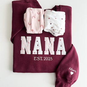 May include: A maroon sweatshirt featuring the embroidered text NANA in block letters with a light pink rainbow pattern, and the text EST. 2025 below. The sleeve displays the name Alara with a heart. Two folded fabric pieces with rainbow and heart prints rest on top.