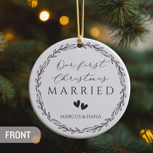 May include: A round white ceramic ornament featuring a black leaf wreath design. The text reads Our first Christmas MARRIED with two small hearts and the names MARCUS & HANA below. The ornament hangs from a gold string.