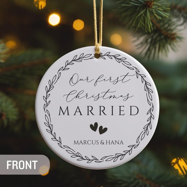 May include: A round white ceramic ornament featuring a black leaf wreath design. The text reads Our first Christmas MARRIED with two small hearts and the names MARCUS & HANA below. The ornament hangs from a gold string.