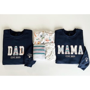 May include: Navy blue sweatshirts, one with "DAD" and the other with "MAMA" in white and blue block letters. Both have "EST. 2025" printed below. A stack of baby blankets in various patterns is placed between the sweatshirts.