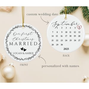 May include: Two round white ceramic ornaments with gold hanging strings. The front features a black leaf wreath, the text Our first Christmas MARRIED, two small hearts, and the names LOGAN & AMBER. The back shows a September 2025 calendar.