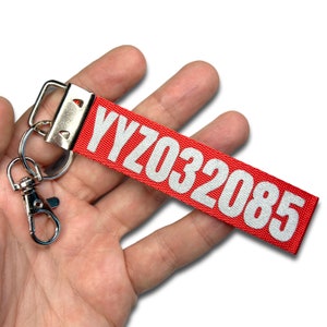 Peut inclure: Un porte-clés rouge avec le texte blanc "YYZ032085". Il est attaché à un mousqueton et un anneau porte-clés en métal argenté. Le porte-clés est tenu dans la main d'une personne.