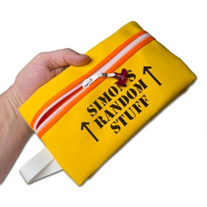 May include: A yellow rectangular pouch with a white zip and orange trim. The text "SIMON'S RANDOM STUFF" is printed in black, with two upward-pointing arrows. A small red charm hangs from the zip.