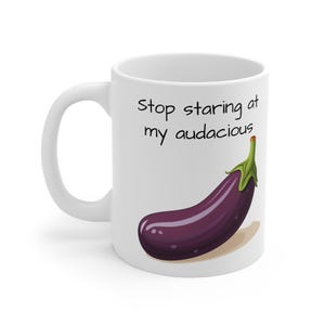 Puede incluir: Taza de cerámica blanca con una ilustración de berenjena morada y el texto "Stop staring at my audacious". La berenjena tiene hojas verdes y una sombra marrón claro.