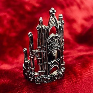 May include: A silver-colored ring with a gothic cathedral design. The ring features intricate details, including arched windows, spires, and a star-shaped emblem. The ring is set against a red background.