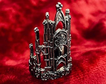 May include: A silver-colored ring with a gothic cathedral design. The ring features intricate details, including arched windows, spires, and a star-shaped emblem. The ring is set against a red background.