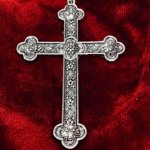 May include: A silver-colored cross pendant with ornate detailing. The cross features a floral design at the center and on the ends of each arm. The pendant is set against a deep red background.