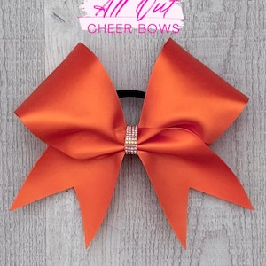 May include: Orange cheer bow with a rhinestone center. The bow is made of satin material and has a black elastic band. The text "All Out Cheer Bows" is in the background.