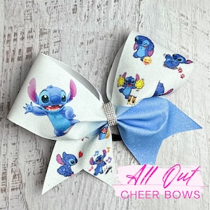 May include: A blue and white cheer bow with a glitter finish. The bow features a cartoon character with blue fur and large ears. The bow is decorated with the text "All Out Cheer Bows".