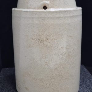 May include: A cylindrical, off-white stoneware crock with a slightly tapered top and a small hole near the top. The surface has a speckled appearance, suggesting an aged or rustic aesthetic. The crock is likely used for food storage or decoration.