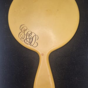 Pu&ograve; includere: Uno specchio a mano vintage giallo pallido, con una testa ovale e un manico abbinato. Lo specchio presenta un monogramma nero ornato sulla testa. L'impugnatura ha la forma di una goccia. Un classico accessorio da toeletta.