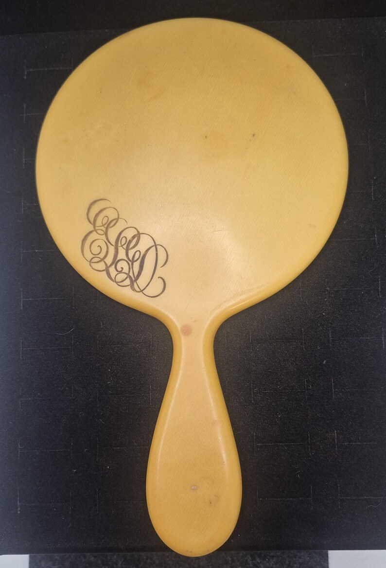Pu&ograve; includere: Uno specchio a mano vintage giallo pallido, con una testa ovale e un manico abbinato. Lo specchio presenta un monogramma nero ornato sulla testa. L'impugnatura ha la forma di una goccia. Un classico accessorio da toeletta.