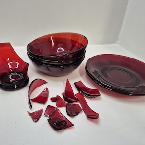 May include: A collection of red glass bowls, plates, and a broken vase. The bowls are stacked, and the plates are side by side. Several shards of red glass are scattered around the other items. The items are on a white surface.