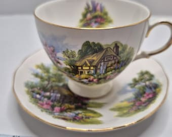 May include: A white teacup and saucer set with a gold rim. The set features a cottage scene with a green roof and a blue sky.