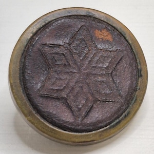 May include: A round, antique-style button with a dark brown center featuring a six-pointed star design. The button has a bronze-colored rim and a textured appearance, suggesting age and wear. The star is deeply etched into the center.