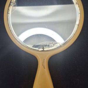 Pu&ograve; includere: Un specchio a mano vintage con una superficie a specchio rotonda, leggermente incrinata. La cornice e il manico sono di colore giallo pallido. Il manico riporta la scritta "Persian Ivory, Tiara, Made in France".