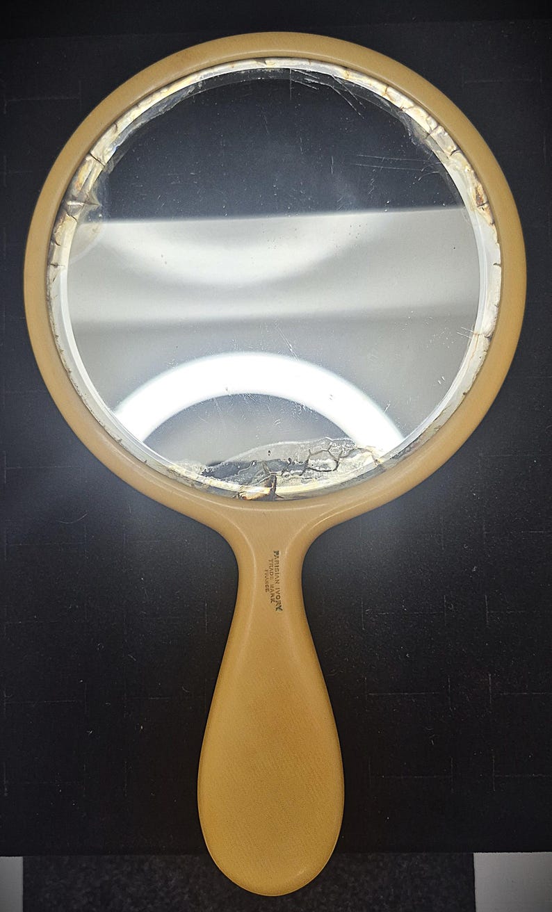 Pu&ograve; includere: Un specchio a mano vintage con una superficie a specchio rotonda, leggermente incrinata. La cornice e il manico sono di colore giallo pallido. Il manico riporta la scritta "Persian Ivory, Tiara, Made in France".