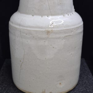 May include: A white ceramic jar with a cylindrical body and a slightly narrower neck. The jar has a crackled glaze and visible age-related wear. The jar is likely an antique or vintage piece, suitable for home decor.
