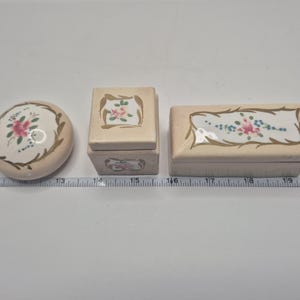 May include: Three vintage ceramic trinket boxes with floral designs. The boxes are in shades of white, cream, and gold. The largest box is rectangular, the middle box is square, and the smallest box is round.