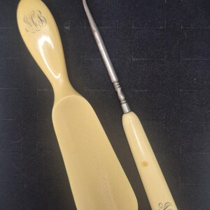 Pu&ograve; includere: Due antichi strumenti color avorio. Uno &egrave; un calzascarpe con un monogramma inciso. L'altro &egrave; un uncinetto in metallo con un manico color avorio, anch'esso con un monogramma inciso. Entrambi gli strumenti sono su una superficie scura.
