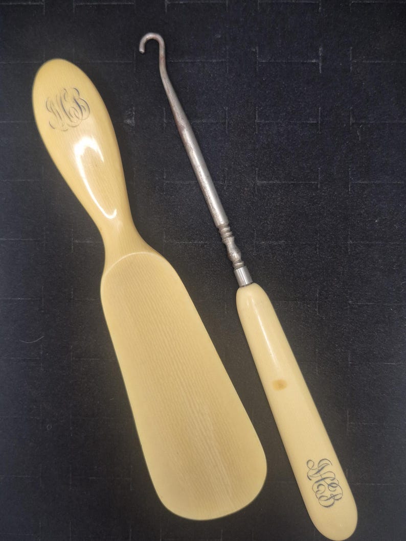 Pu&ograve; includere: Due antichi strumenti color avorio. Uno &egrave; un calzascarpe con un monogramma inciso. L'altro &egrave; un uncinetto in metallo con un manico color avorio, anch'esso con un monogramma inciso. Entrambi gli strumenti sono su una superficie scura.