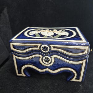 May include: A square, ceramic jewelry box with a dark blue glaze and white floral design on the lid. The box has a white trim and decorative floral accents on the front. The box is approximately 4 inches wide and 4 inches long.