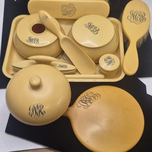 May include: A vintage vanity set in a creamy yellow hue, featuring a hand mirror, brushes, and various cosmetic containers. Each piece is adorned with elegant black monogrammed initials. The set includes a tray, a powder jar, and a small container.