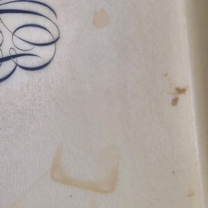 Pu&ograve; includere: Primo piano di una superficie color crema con scritte in corsivo blu scuro nell'angolo in alto a sinistra. Sono visibili diverse macchie marroni sulla superficie, tra cui una macchia quadrata.