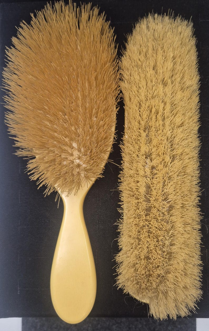 Pu&ograve; includere: Due spazzole per capelli antiche. Una &egrave; ovale con un lungo manico curvo. L'altra &egrave; rettangolare. Entrambe hanno setole marrone chiaro e manici color crema. Le spazzole sono probabilmente realizzate con materiali naturali.