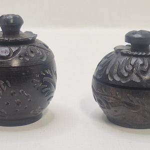 May include: Two dark gray, round lidded jars with carved floral designs. The jars have a spherical shape with a small knob on top of the lid. The jars are made of a dark gray material.