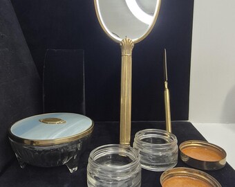 Può includere: Un set da toeletta vintage con uno specchio a mano, un piccolo barattolo e due piccoli contenitori con bordo dorato. Lo specchio a mano ha un manico e una cornice dorati. Il set include un contenitore blu e oro con coperchio e uno strumento dorato.