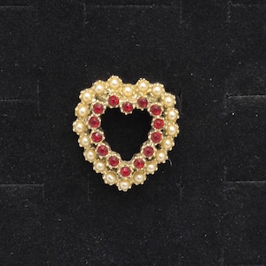 May include: A heart-shaped brooch with a gold-tone frame. The brooch is adorned with small, round, faux pearls and red, round gemstones. The heart shape is open in the center, and the brooch is set against a black background.