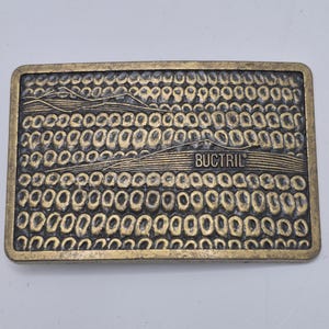 May include: A gold-toned metal belt buckle with a textured design of corn stalks and the word "Buc-Tri" in black lettering.