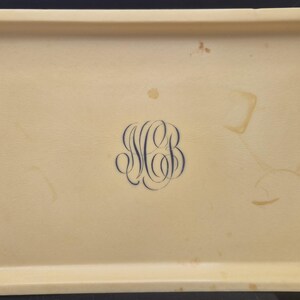 Pu&ograve; includere: Vassoio rettangolare beige con angoli arrotondati. Un monogramma blu scuro con le lettere "MB" &egrave; centrato sul vassoio. Il vassoio presenta alcune macchie.