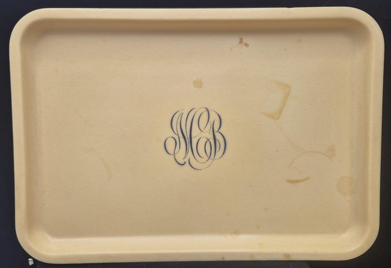 Pu&ograve; includere: Vassoio rettangolare beige con angoli arrotondati. Un monogramma blu scuro con le lettere "MB" &egrave; centrato sul vassoio. Il vassoio presenta alcune macchie.