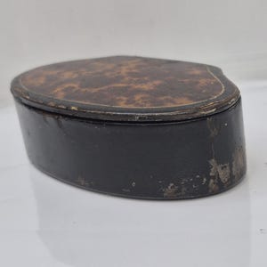 May include: An oval-shaped box with a dark brown, mottled, burl-like pattern on the lid and solid black sides. The lid features a thin, light-colored decorative border line. The black base shows signs of wear with visible surface scuffing.
