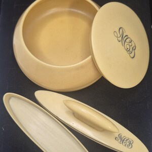 Pu&ograve; includere: Un set da toeletta vintage color crema, con una ciotola rotonda con coperchio e due applicatori ovali. Il coperchio e un applicatore sono incisi con le iniziali "MCB" in un carattere corsivo. Il set &egrave; probabilmente realizzato in un materiale liscio, forse simile all'avorio.