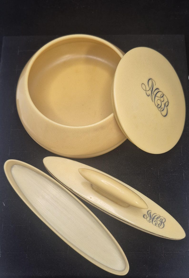 Pu&ograve; includere: Un set da toeletta vintage color crema, con una ciotola rotonda con coperchio e due applicatori ovali. Il coperchio e un applicatore sono incisi con le iniziali "MCB" in un carattere corsivo. Il set &egrave; probabilmente realizzato in un materiale liscio, forse simile all'avorio.