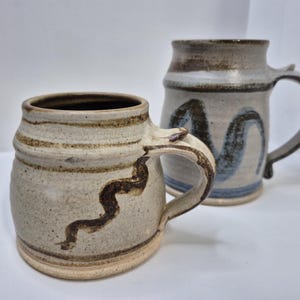 May include: Two handmade ceramic mugs. The mug in the foreground is beige with brown stripes and a dark brown, wavy design. The second mug is light gray with blue and brown brushstrokes. Both mugs have large handles.