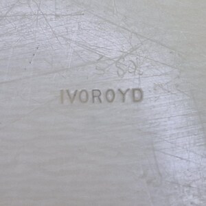 Pu&ograve; includere: Primo piano di una superficie colorata chiara con la parola "IVOROYD" in rilievo al centro. La superficie ha un aspetto strutturato con linee sottili e graffi.