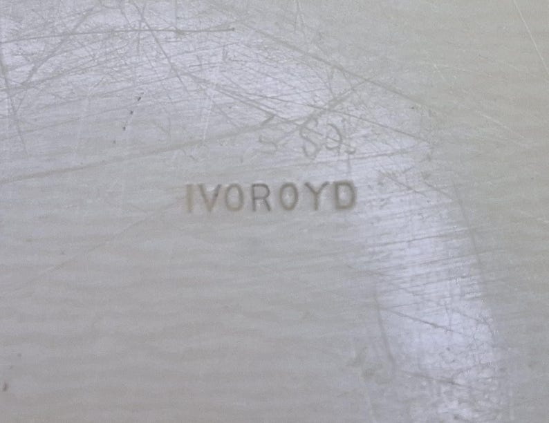 Pu&ograve; includere: Primo piano di una superficie colorata chiara con la parola "IVOROYD" in rilievo al centro. La superficie ha un aspetto strutturato con linee sottili e graffi.
