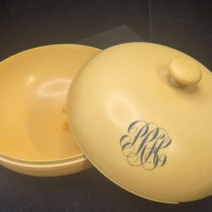 Pu&ograve; includere: Un contenitore rotondo giallo pallido vintage con coperchio abbinato. Il coperchio presenta un piccolo pomello e un monogramma blu. Il contenitore &egrave; aperto, rivelando l'interno. Il design suggerisce un oggetto decorativo o di stoccaggio.