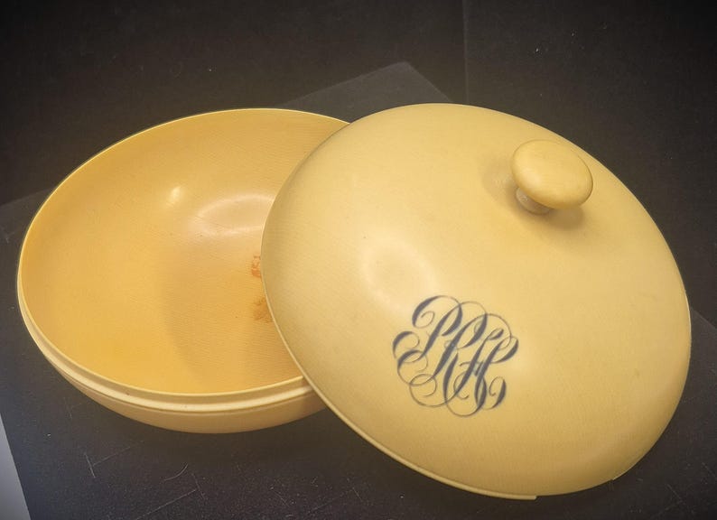 Pu&ograve; includere: Un contenitore rotondo giallo pallido vintage con coperchio abbinato. Il coperchio presenta un piccolo pomello e un monogramma blu. Il contenitore &egrave; aperto, rivelando l'interno. Il design suggerisce un oggetto decorativo o di stoccaggio.
