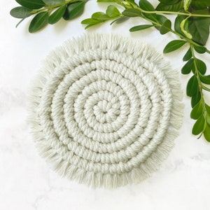 May include: A round, cream-colored coaster with a fringe border. The coaster is made of woven rope and has a circular pattern.