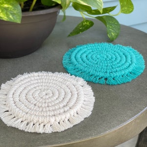 May include: Two round crocheted coasters with fringe. One coaster is white and the other is turquoise.
