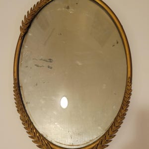 Könnte beinhalten: Ein ovaler Wandspiegel mit einem goldfarbenen Metallrahmen, der an den Seiten mit geprägten Lorbeerblattmustern verziert ist. Am unteren Rand des Rahmens befindet sich ein kleines, mittiges schleifenartiges Detail mit undeutlichem Text.
