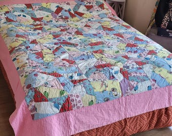 May include: A patchwork quilt with a pink border and a brown bed skirt. The quilt is made of various fabrics in shades of blue, pink, yellow, and red.