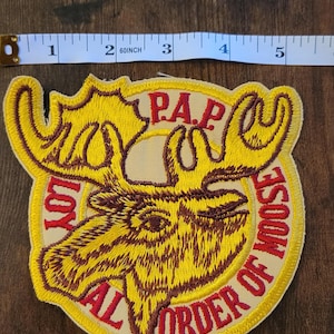 May include: Embroidered patch featuring a moose head with large antlers. The patch is yellow with brown and red accents. Text includes "LOYAL ORDER OF MOOSE" and "P.A.P.". The patch measures approximately 10 cm in diameter.