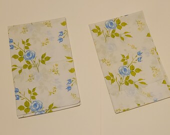 May include: Two rectangular white fabric napkins featuring a repeating floral pattern of light blue roses and green leaves. The design includes small yellow sprigs scattered across the white background, creating a consistent botanical print on both items.